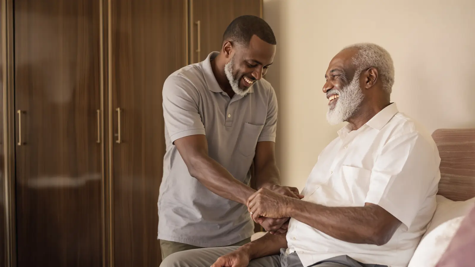 A caregiver sitting with an elderly Nigerian man on the edge of his bed at home, both smiling