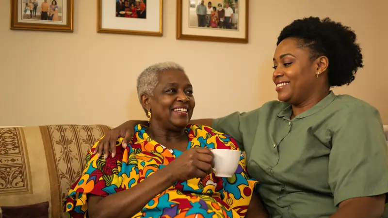 An elderly Nigerian woman with a cup of tea on the sofa in the evening as her daughter comforts her